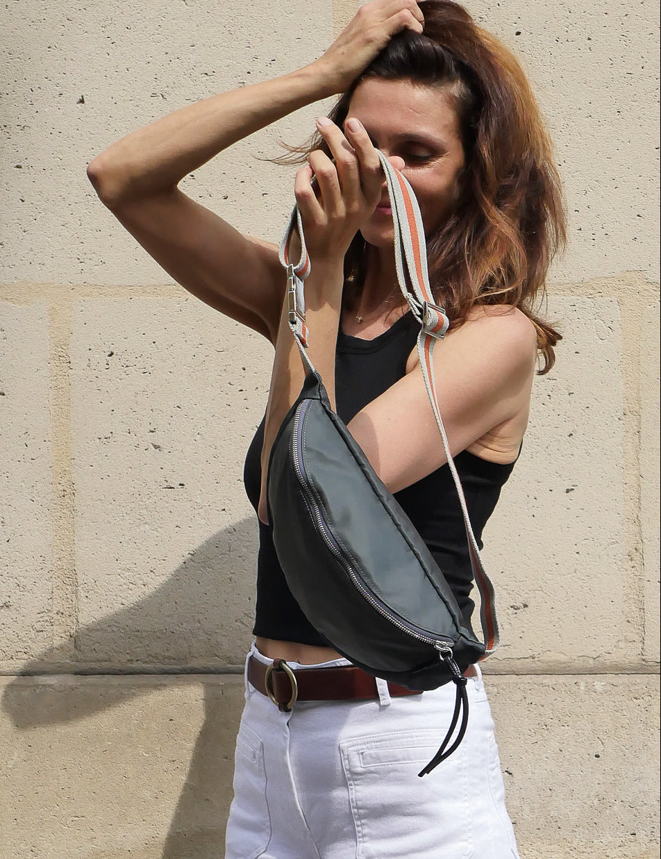 Dark grey satin nylon fanny pack designed and made in Paris with a striped  adjustable crossbody strap, worn by a Parisian beauty on the streets of Paris.