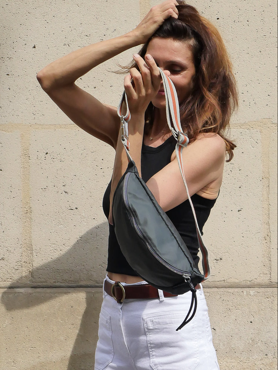 Dark grey satin nylon fanny pack designed and made in Paris with a striped  adjustable crossbody strap, worn by a Parisian beauty on the streets of Paris.