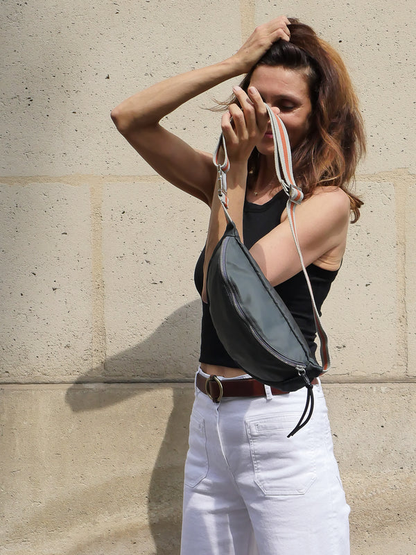 Dark grey satin nylon fanny pack designed and made in Paris with a striped  adjustable crossbody strap, worn by a Parisian beauty on the streets of Paris.